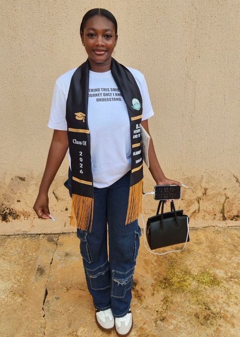 The image features a young woman wearing a graduation cap and gown with a black t-shirt and blue jeans. She has a black purse slung over her shoulder and a cell phone in her hand, suggesting she might be checking messages or making plans for the next day. The background of the photo is a plain wall, which further emphasizes the focus on the woman and her graduation attire.