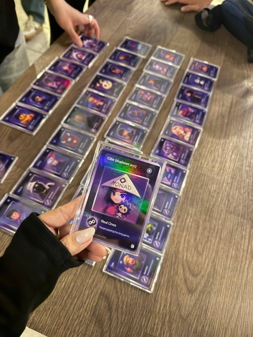 A hand holds up a holographic 'Monad' trading card labeled 'Root Cross' with a Road Map series, displaying rows of similar cards laid out on a wooden table.