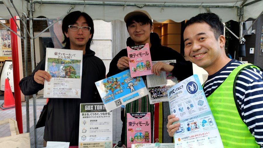 Three smiling Japanese men standing under a tent holding up various informational flyers and brochures about 東ティモール (East Timor) and other topics, including a GOX TOKYO pamphlet.
