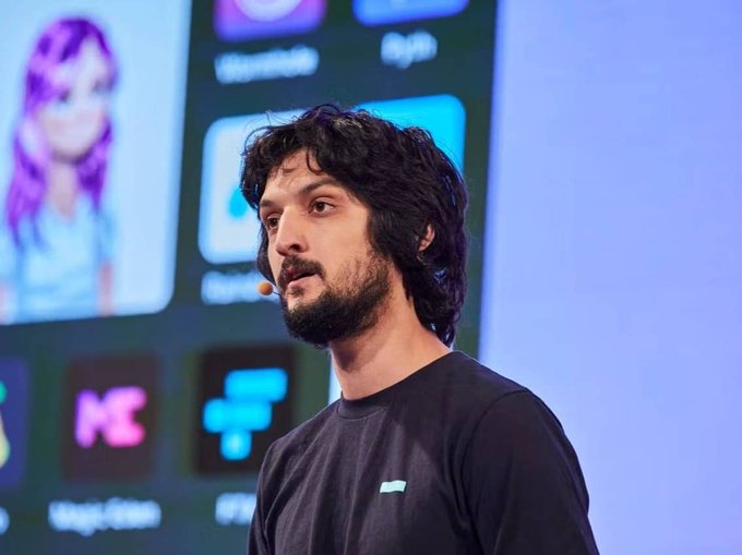 A bearded man with dark curly hair speaking on stage with a microphone headset, wearing a dark sweatshirt, with app icons visible on a screen behind him.