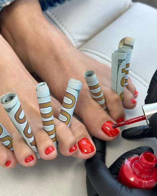 A close-up of a pedicure in progress, showing toenails being painted with bright red polish while paper nail forms are wrapped around the toes for extensions.
