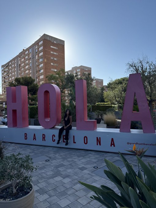 Woman sitting on a large pink 'HOLA BARCELONA' sign in a plaza with palm trees, plants, and apartment buildings in the background under a sunny sky.