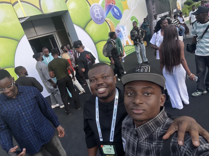 Two young men smile for a selfie at what appears to be a cryptocurrency/NFT event, with one wearing a lanyard and a cap, while a crowd gathers near a building decorated with crypto-themed murals featuring Bitcoin, Ethereum, and Tron logos. The image connects to the post by showing the friends mentioned—@NFT__NG, @CryptoDefiLord, and @teddi_speaks—at the event.