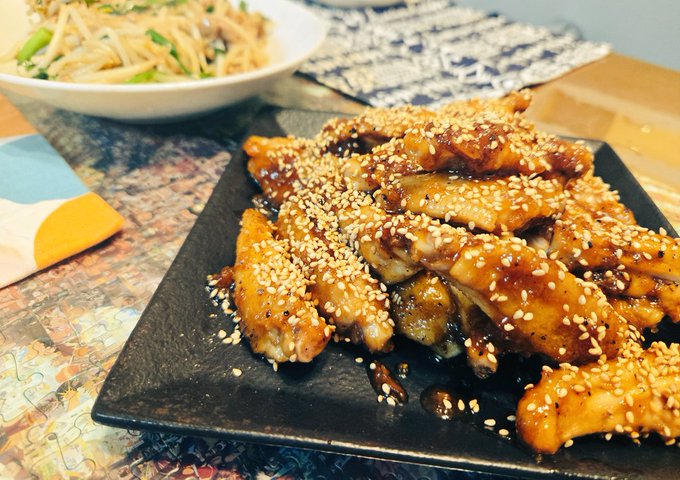The image features a plate of food with sesame seeds sprinkled over it, likely chicken wings or another type of meat dish. The plate is placed on top of a table next to a bowl filled with noodles. In the background, there are some other dishes as well. The photo appears to be related to a cooking tutorial or recipe, possibly for preparing a meal that includes both chicken and noodles.
