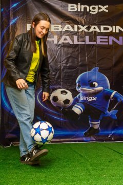A photo of a woman in a black jacket and yellow shirt posing next to a BingX mascot character holding a soccer ball, with a 'BingX' and 'BAIXADIN HALLEN' banner in the background.