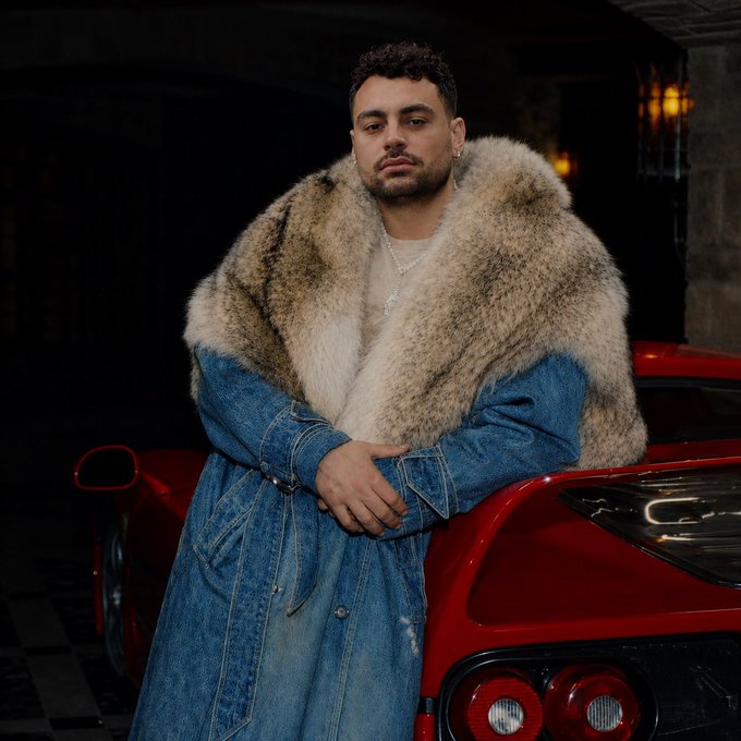 A man with short curly hair poses in a denim coat with a large fur collar, leaning against a red sports car in a dimly lit setting.