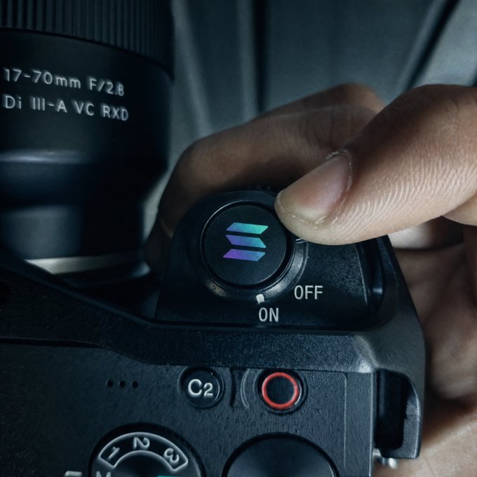 A close-up of a camera with a Tamron 17-70mm F/2.8 Di III-A VC RXD lens, showing a finger near the power switch with the Solana logo engraved on the camera body.