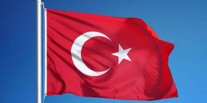 The image features a red flag flying high against a blue sky with white clouds. The flag has a crescent moon design at its center, which is the Turkish national symbol. The flag appears to be waving in the wind, indicating that it's blowing gently. In the background, there are some texts that read "BREAKING: Ripple is expanding the reach of its dollar-backed stablecoin, #RLUSD, into Turkey". This suggests that the image might have been taken during a time when Ripple was actively promoting its RLUSD token in Turkey or discussing its potential use in the country.