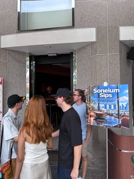 A group of people gathered outside an entrance, looking at a digital billboard displaying 'Soneium Gigs' in the Tokyo Shibuya area.