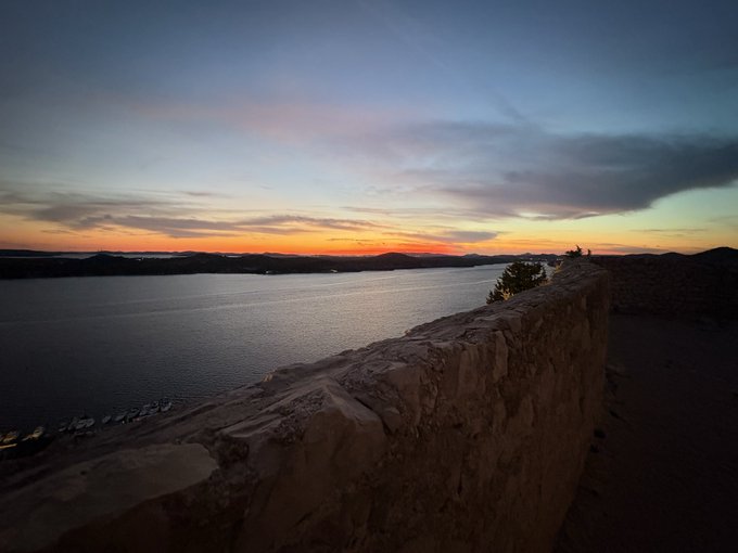A scenic landscape photograph showing a sunset over a large body of water with mountains in the distance, taken from what appears to be an elevated stone structure or wall.