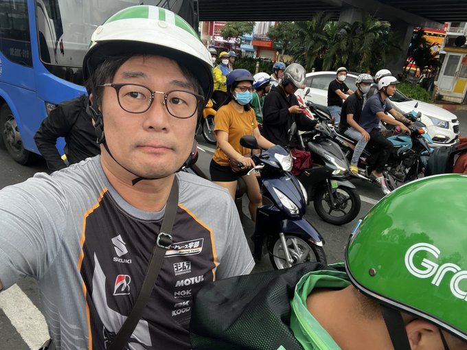 A man in a Suzuki branded shirt and helmet is caught in traffic in Vietnam, likely while navigating the challenges of ASEAN marketing for Matsuya Ginza, suggesting a need for remittance solutions like JPYC. The "Grab" logo on another helmet further emphasizes the transportation context.