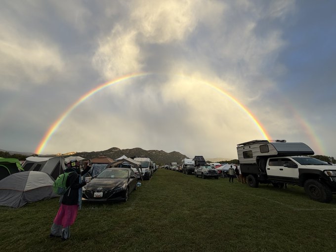 A wide shot of a grassy campsite filled with tents, cars, and camper trucks beneath a dramatic sky featuring a large, complete rainbow arching across the clouds.