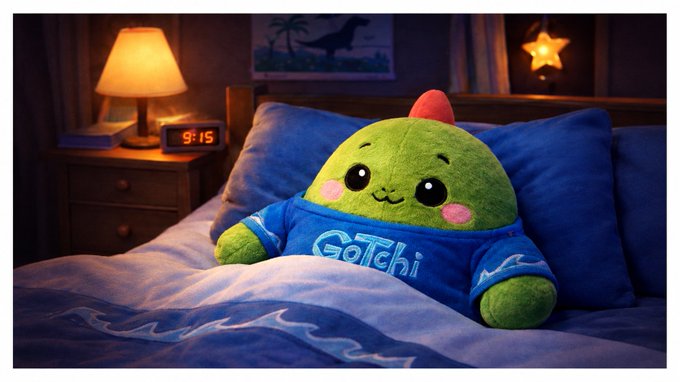 An image of a green Tamagotchi-style plush toy wearing a blue 'Gotchi' shirt, sitting in a blue bed next to a nightstand with a lamp and a digital clock reading '9:15'.