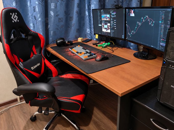 A gaming and trading setup featuring a black and red Sharkoon gaming chair, a mechanical keyboard, a mouse, and dual monitors displaying trading charts and chat interfaces.