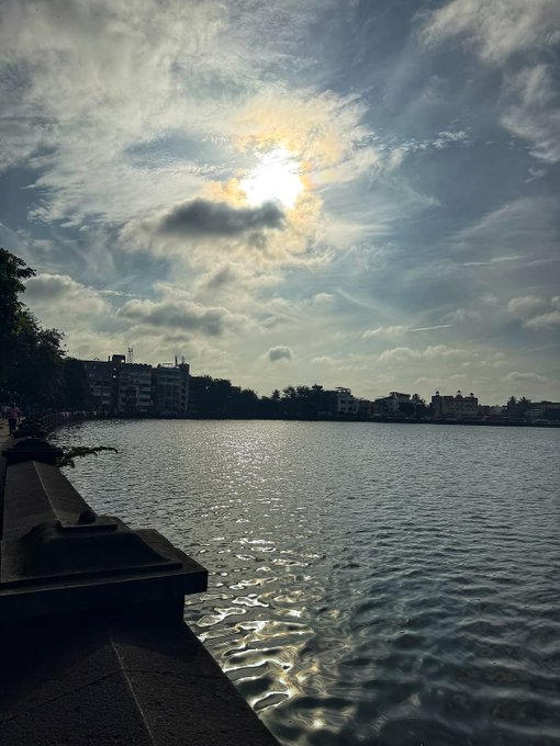 A serene morning view of a calm waterway with the sun breaking through scattered clouds, casting shimmering reflections on the surface, while a stone railing and distant cityscape frame the peaceful scene. The tranquil sunrise perfectly complements the fresh, optimistic start to the new week, embodying the "new week, new wins" energy.