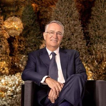 Ricardo Salinas, a Mexican billionaire businessman, smiles warmly while seated in front of a backdrop of illuminated greenery, accompanying a post discussing his bold prediction that Bitcoin could reach $1.86 million per coin to match gold's market capitalization.