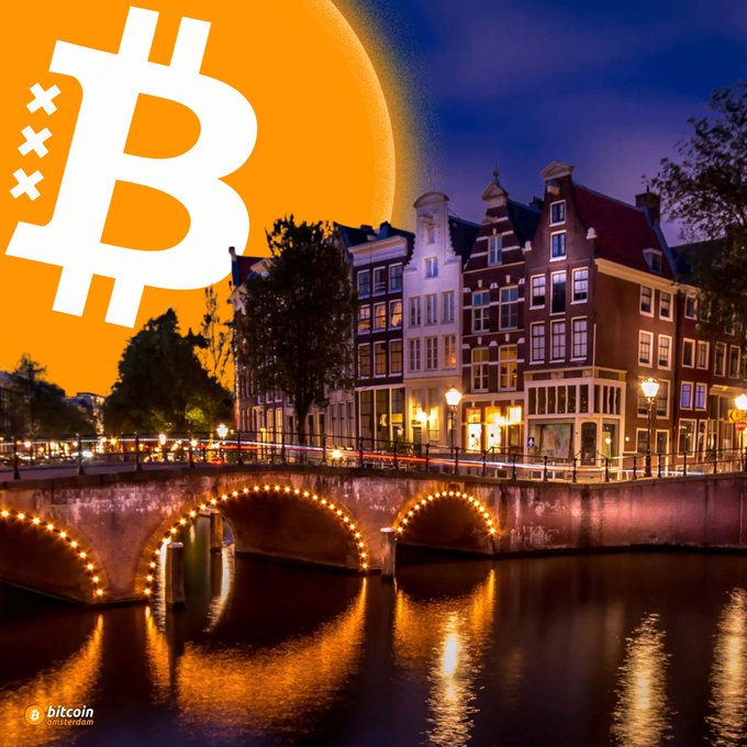 The image features the iconic Amsterdam canal houses and a lit stone bridge reflected in the water at dusk, with a large glowing orange Bitcoin logo overlaid in the sky and the "Bitcoin Amsterdam" logo in the corner. The scene visually promotes the upcoming Bitcoin Amsterdam event, using the city's recognizable architecture to build excitement for the gathering of Bitcoiners in just 60 days.