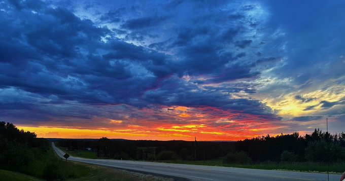 A vibrant sunset paints the sky with orange, pink, and deep blue clouds over a quiet country road lined with trees.