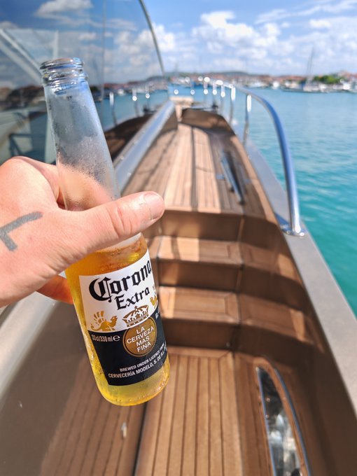 A hand holds a chilled bottle of Corona Extra beer in the foreground, with a wooden boat deck and turquoise Caribbean waters visible in the background.