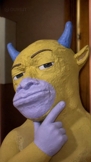 A claymation-style sculpture of a yellow creature with blue horns and a purple snout, pointing at its face with a purple hand, with an 'OURBIT' watermark in the top left.