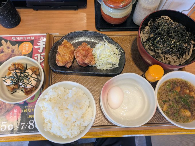 A Japanese lunch set featuring a bowl of white rice, soba noodles topped with seaweed, fried chicken with shredded cabbage, a raw egg, miso soup, and a small side dish, with a promotional flyer visible showing "期間限定価格" and "7/8" dates. The meal pairs with the post's humor about taking a lunch break from STEPN/GO, missing the session window, and overeating ("ご飯を大盛り" and "蕎麦を2倍盛り") as a result—ironically enjoying a hearty meal while lamenting needing to be more mindful next time.