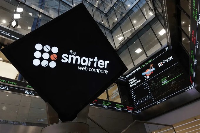 The image shows a digital display inside a financial building (likely the London Stock Exchange) featuring "the smarter web company" logo prominently on a black screen, alongside stock tickers including "LM.L" and "EDV.L," with additional screens displaying "THE SMARTER WEB" and references to Bitcoin and a "Digital, Capital, Delighted" campaign—visually tying to the company's announcement of floating perpetual preferred shares on the LSE under ticker MORE to fund Bitcoin acquisitions.