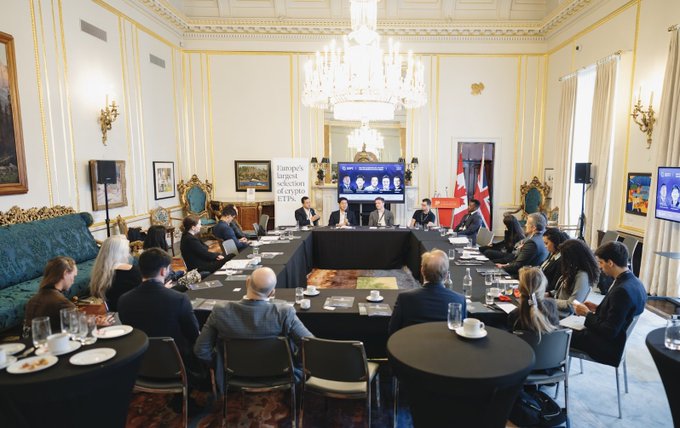 The image shows a group of people sitting around a large conference table, engaged in a meeting or presentation. There are at least 12 individuals visible in the scene, some wearing ties, suggesting a formal gathering. The room has a grand appearance with white walls and gold accents, giving it an elegant atmosphere. A TV screen is mounted on one wall, possibly displaying relevant information for the discussion.

The people appear to be listening attentively as they participate in what seems to be a business meeting or presentation. There are several chairs positioned around the table, accommodating the attendees. The room also features multiple cups and wine glasses placed strategically throughout the space, indicating that refreshments have been provided during the event.
