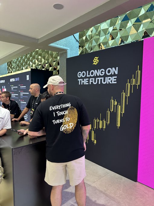 A man wearing a black t-shirt reading 'EVERYTHING I TOUCH TURNS TO GOLD' stands at a crypto conference booth with signage 'GO LONG ON THE FUTURE' and a gold candlestick chart.