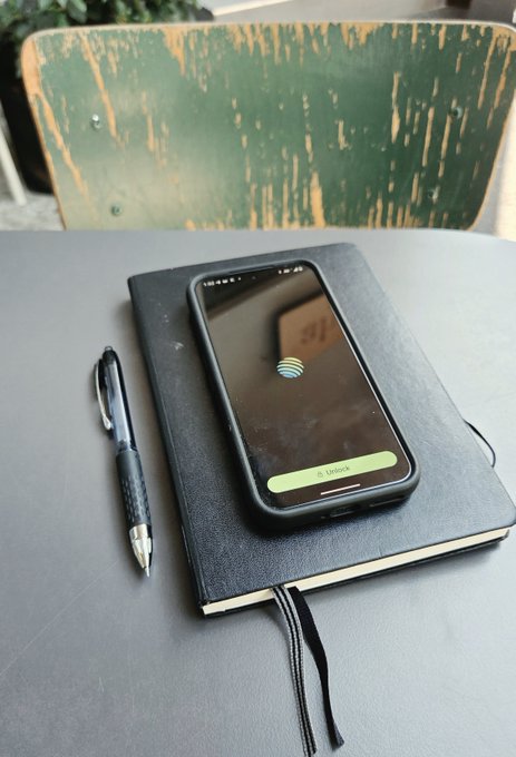A black smartphone displaying an AT&T lock screen rests on a black notebook with a bookmark, next to a black pen, all sitting on a grey table in front of a distressed green chair.