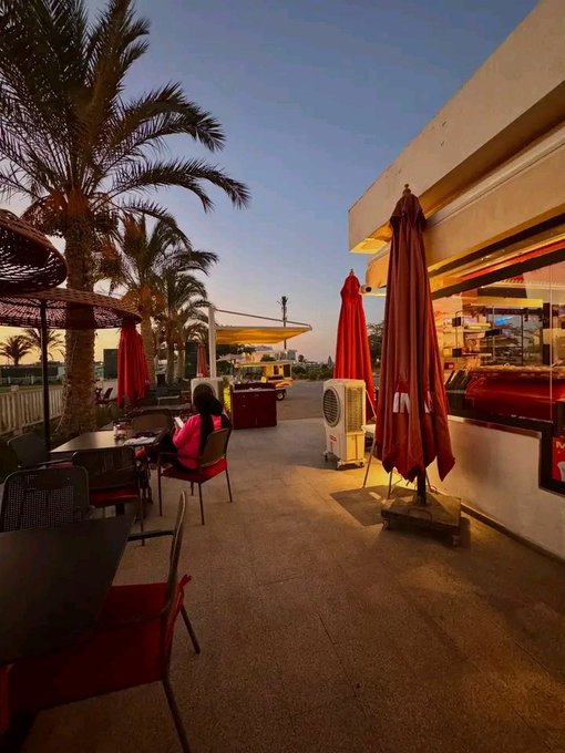 An outdoor café or restaurant patio at sunset, featuring palm trees, red umbrellas, tables with chairs, and a person seated.