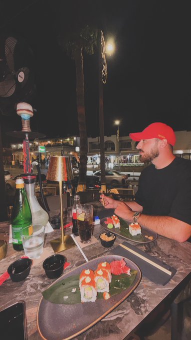 A man wearing a red baseball cap and black shirt dines outdoors at night, enjoying a spread of sushi rolls served on green leaf-lined plates, accompanied by chopsticks, a cocktail, and a sparkling water bottle, all illuminated by a table lamp with a palm tree in the background—setting a relaxed evening scene that fits the casual, friendly vibe of a "Gm friends 🖤" greeting.