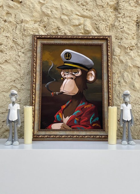 A framed cartoon-style painting of a monkey wearing a white captain's hat and smoking a cigar, displayed on a shelf between two white candles and flanked by small grey monkey figurines.
