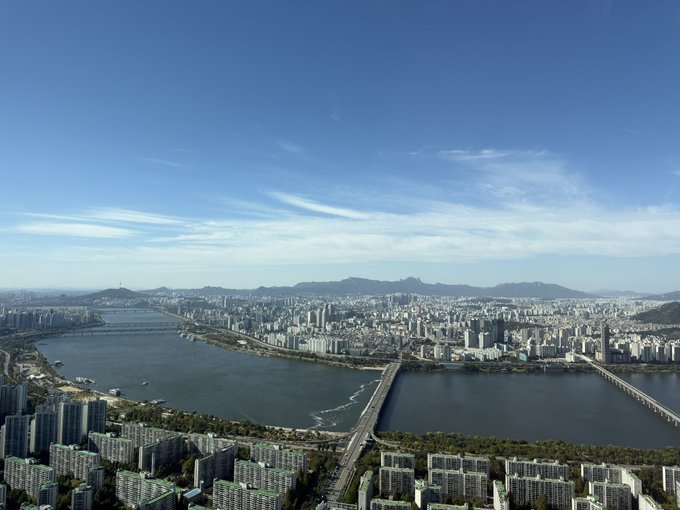 An aerial daytime view of a sprawling cityscape with a wide river running through it, multiple bridges crossing the water, and apartment buildings lining both banks under a blue sky.