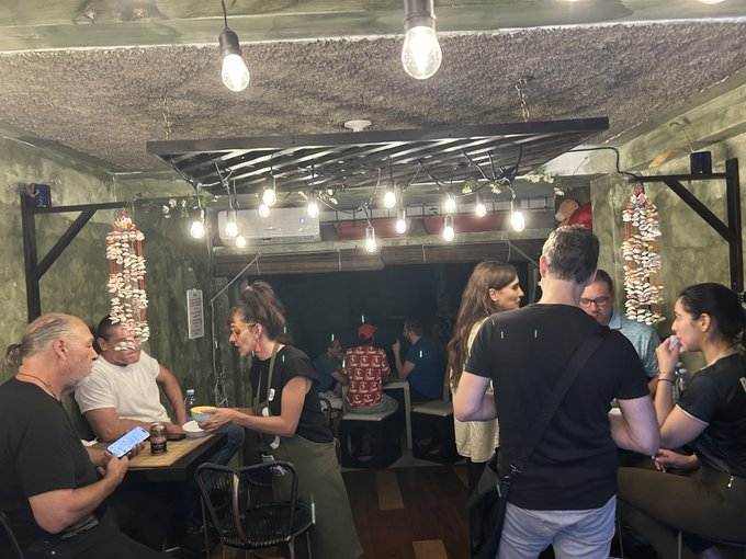 People mingle in a dimly lit venue with exposed concrete, string lights, and hanging decorations, with one person looking at a smartphone.