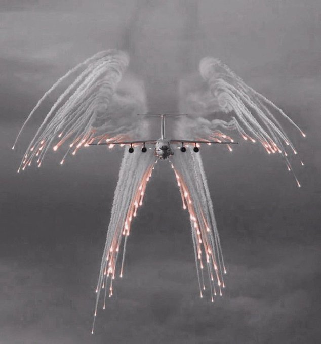 A military transport aircraft deploys a massive display of flares from its wingtips and fuselage against a dark, cloudy sky.