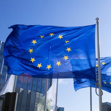 An EU flag billows against a blue sky, symbolizing the European Central Bank's launch of Pontes—a new initiative enabling institutions to settle tokenized asset trades using central bank money rather than stablecoins, with full rollout anticipated by 2028.