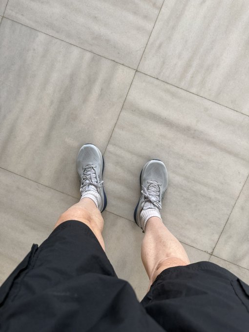 A first-person perspective photo looking down at a person's legs wearing black shorts, white socks, and gray athletic sneakers standing on a tiled floor.