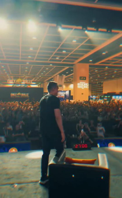 A man stands on a stage facing a large audience at an indoor event called 'Crazy Bulls,' with a timer showing 12:00 visible in the background.