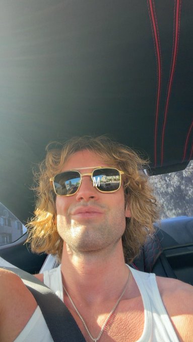A man with long curly blonde hair and aviator sunglasses takes a selfie from inside a convertible, wearing a white tank top and silver chain. The image complements the caption by playfully contrasting his real-life "cool guy" appearance with the casual, ironic tone of embracing his "cartoon profile picture crypto internet friends."