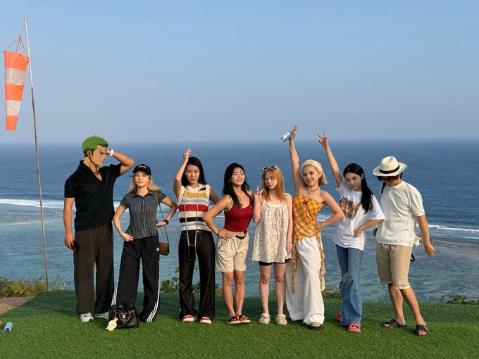 Seven young adults pose together on a grassy cliffside overlooking the ocean, with a green-haired person on the left making peace signs alongside others in casual summer attire.