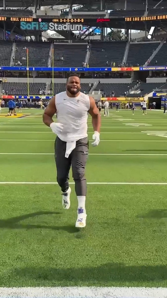 A muscular man in a white sleeveless shirt and gray pants walking on the field at SoFi Stadium, with the Los Angeles Rams visible in the background signage.