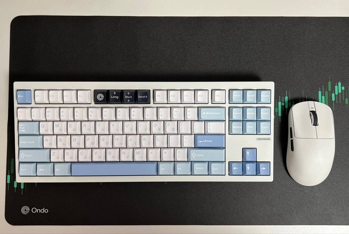 A blue and white mechanical keyboard sits on a black 'Ondo' desk mat with green candlestick graphics, next to a white wireless mouse.
