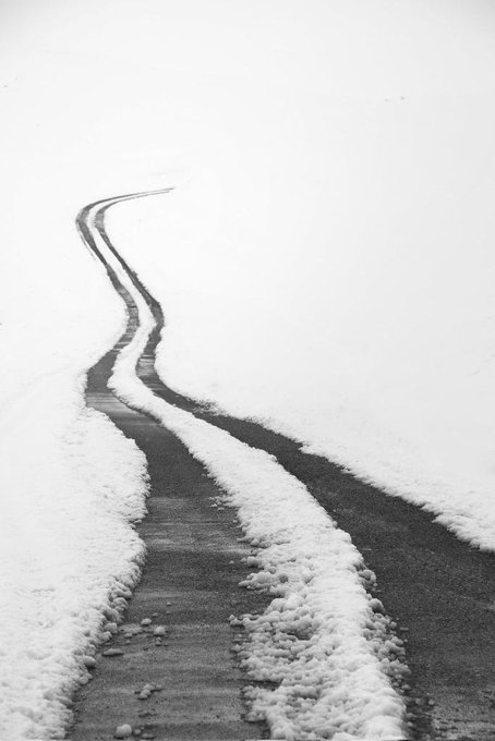 A black-and-white photo of a winding path or road cutting through a snow-covered landscape, with tire tracks visible on the cleared surface.