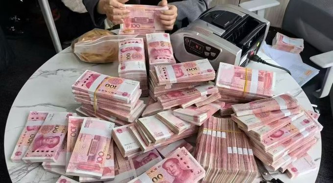 The image shows a person sitting at a table with stacks of 500U Chinese currency notes, possibly from China or Hong Kong. The person appears to be holding one of these stacks of money, possibly preparing for an event or transaction involving large amounts of cash. The scene suggests that the individual is involved in some financial activity or has recently received a significant sum of money.