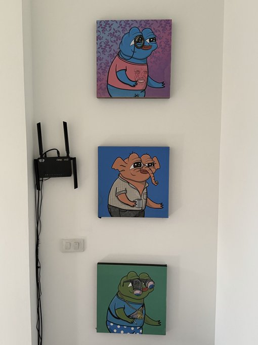 Three canvases hang vertically on a white wall, featuring cartoon-style portraits of a blue bird in a pink shirt, an elephant in a checkered shirt, and a crocodile in a blue shirt; a black ASUS router with two antennas sits on the wall to the left.
