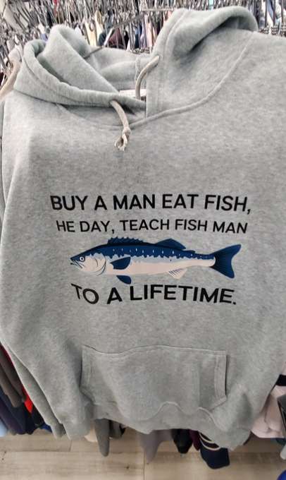 A gray hoodie hanging on a rack featuring a blue fish graphic and the text "BUY A MAN EAT FISH, HE DAY, TEACH FISH MAN TO A LIFETIME."
