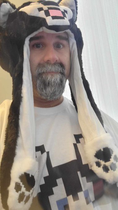 A man wearing a plush animal-themed hat with ears and paw mittens, featuring a pixelated face design, looking at the camera.