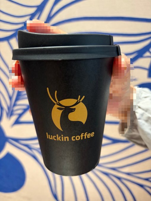A black takeaway cup from Luckin Coffee featuring its signature golden deer logo, held by a hand against a blue and white patterned background.