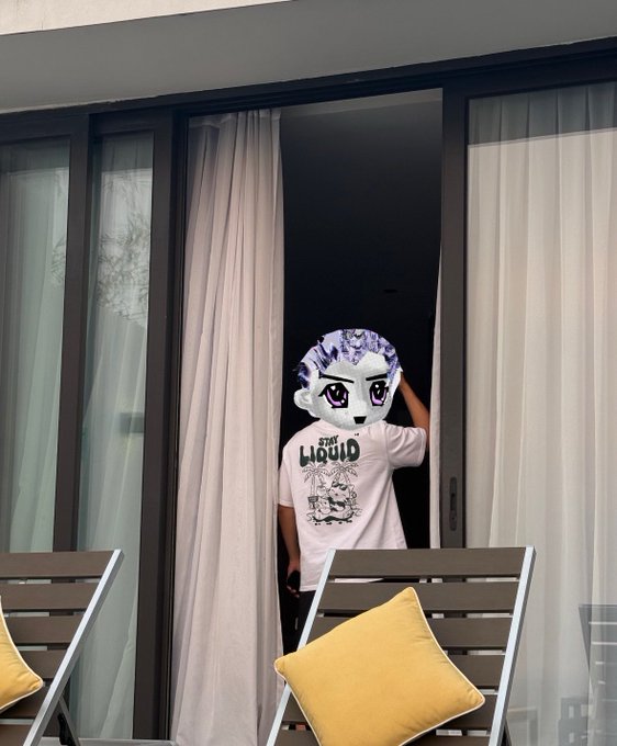 A person wearing a white graphic t-shirt standing on a balcony behind a window, wearing a large mascot-style head mask of a character with purple hair and purple eyes.