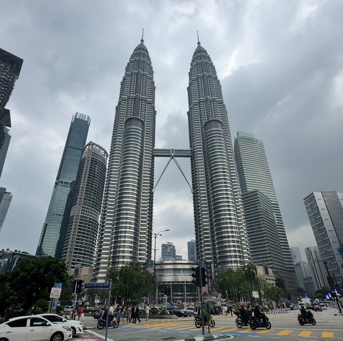 The iconic Petronas Twin Towers in Kuala Lumpur, Malaysia, viewed from street level under a cloudy sky with cars and motorcycles in the foreground.